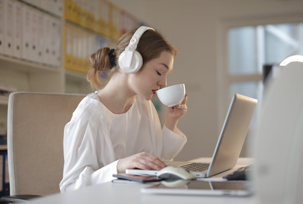 Woman wearing headphones and sipping coffee while working on a laptop at a desk.